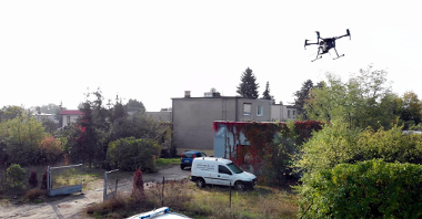 Zdjęcie przedstawia budynki jednorodzinne. Na podwórku jednego z nich, który znajduje się najbliżej stoją dwa samochody. W powietrzu unosi się dron.