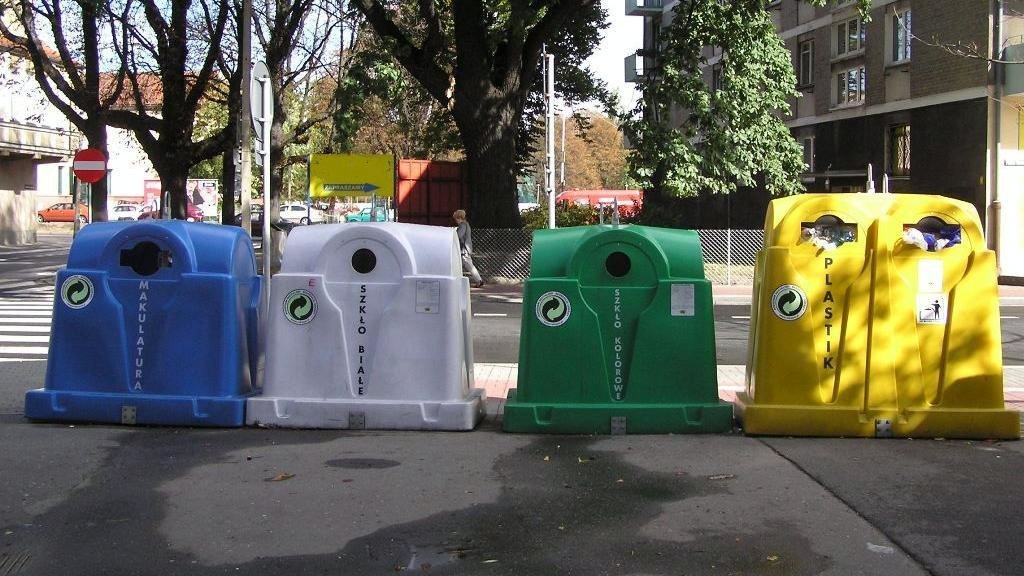 Na zdjęciu cztery różnokolorowe pojemniki na odpady stojące w rzędzie obok siebie - grafika artykułu