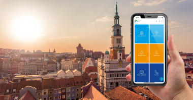 Zdjęcie przedstawia dłoń trzymającą smartfon z działającą aplikacją Poznań Smart City. Aplikacja wyświetla widok menu głównego. W tle widoczny jest Stary Rynek wraz z otaczającymi go kamienicami. Sceneria utrzymana jest w świetle zachodzącego słońca.