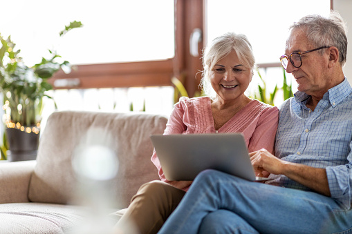 Na fotografii widać parę uśmiechniętych seniorów, kobietę i mężczyznę, którzy korzystają z rozwiązań online. Pracują przed komputerem, uśmiechnięci, razem. - grafika artykułu
