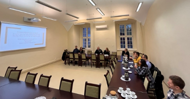 Zdjęcie sali, na której odbywa się zebranie. Kilka osób siedzi przy stołach, na których znajduje się kawa. Na jednej ze ścian projektor wyswietla prezentację.