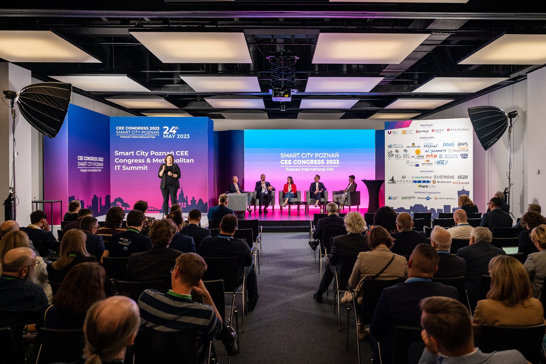 Zdjęcie panelu dyskusyjnego na konferencji na pierwszym planie widownia, na drugim planie scena z panelistami. - grafika artykułu