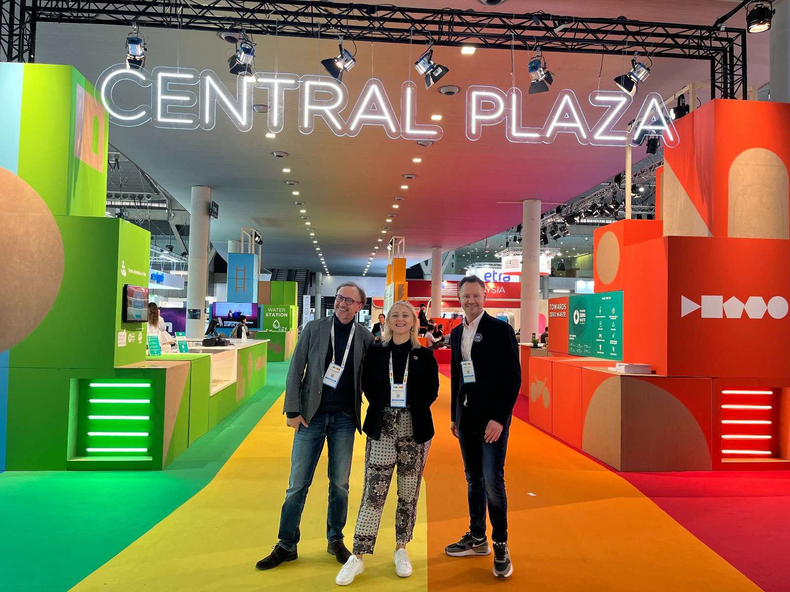 Zdjęcie przedstawia trzy osoby stojące i uśmiechające się na tle kolorowej ekspozycji - grafika artykułu