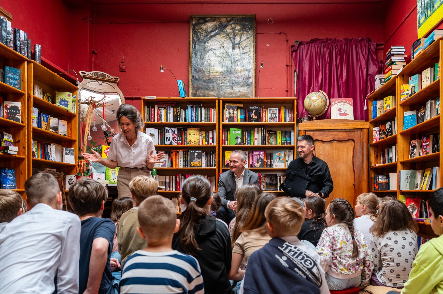 Zdjęcie przedstawia dzieci siedzących na dywanie pośród półek z książkami. W pomieszczeniu znajdują się też dwaj dorośli mężczyźni i kobieta. - grafika artykułu