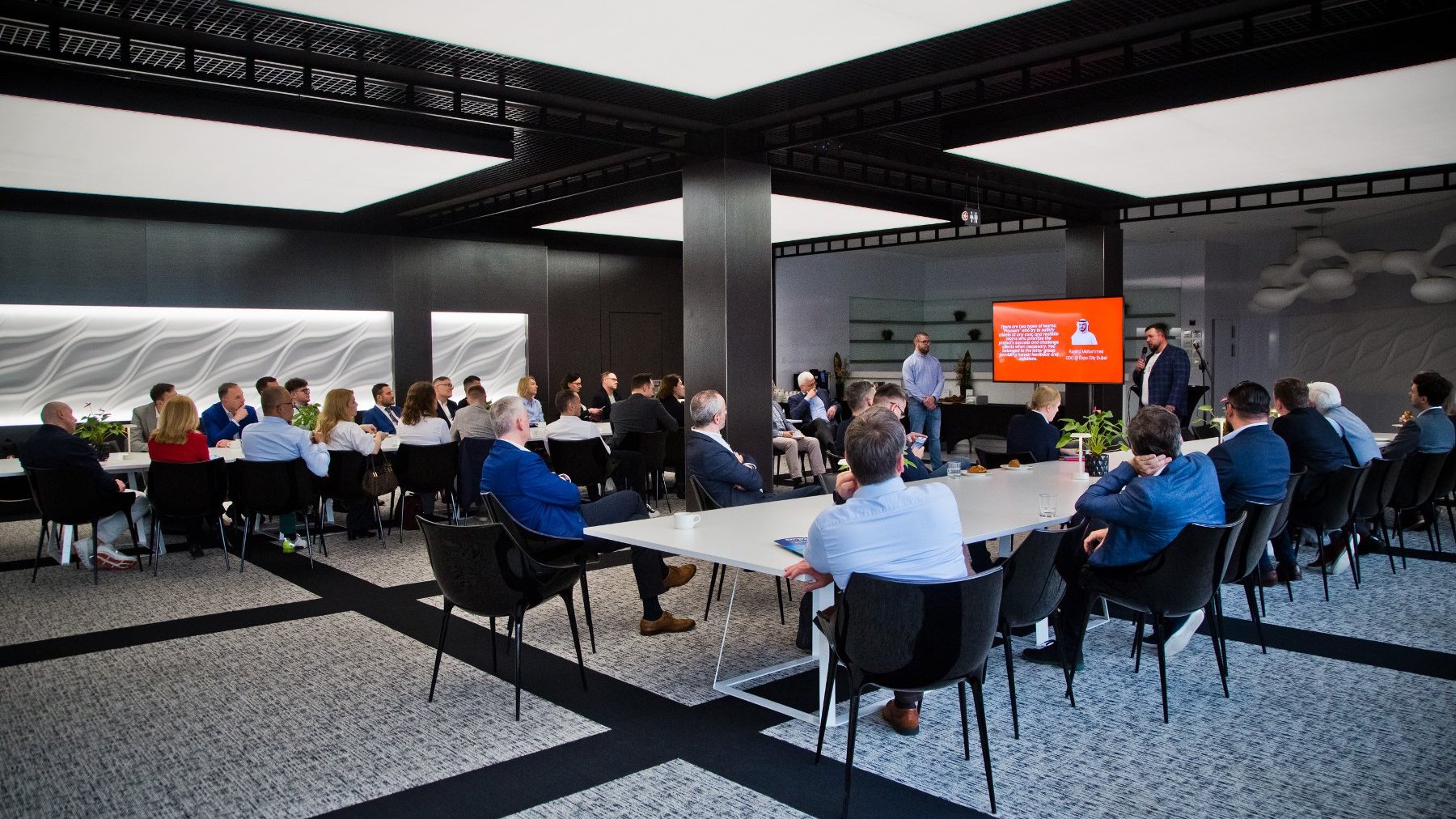 Sala konferencyjna, przy stołach siedzą ludzie, przed nimi ekran, na której wyświetlana jest prezentacja.