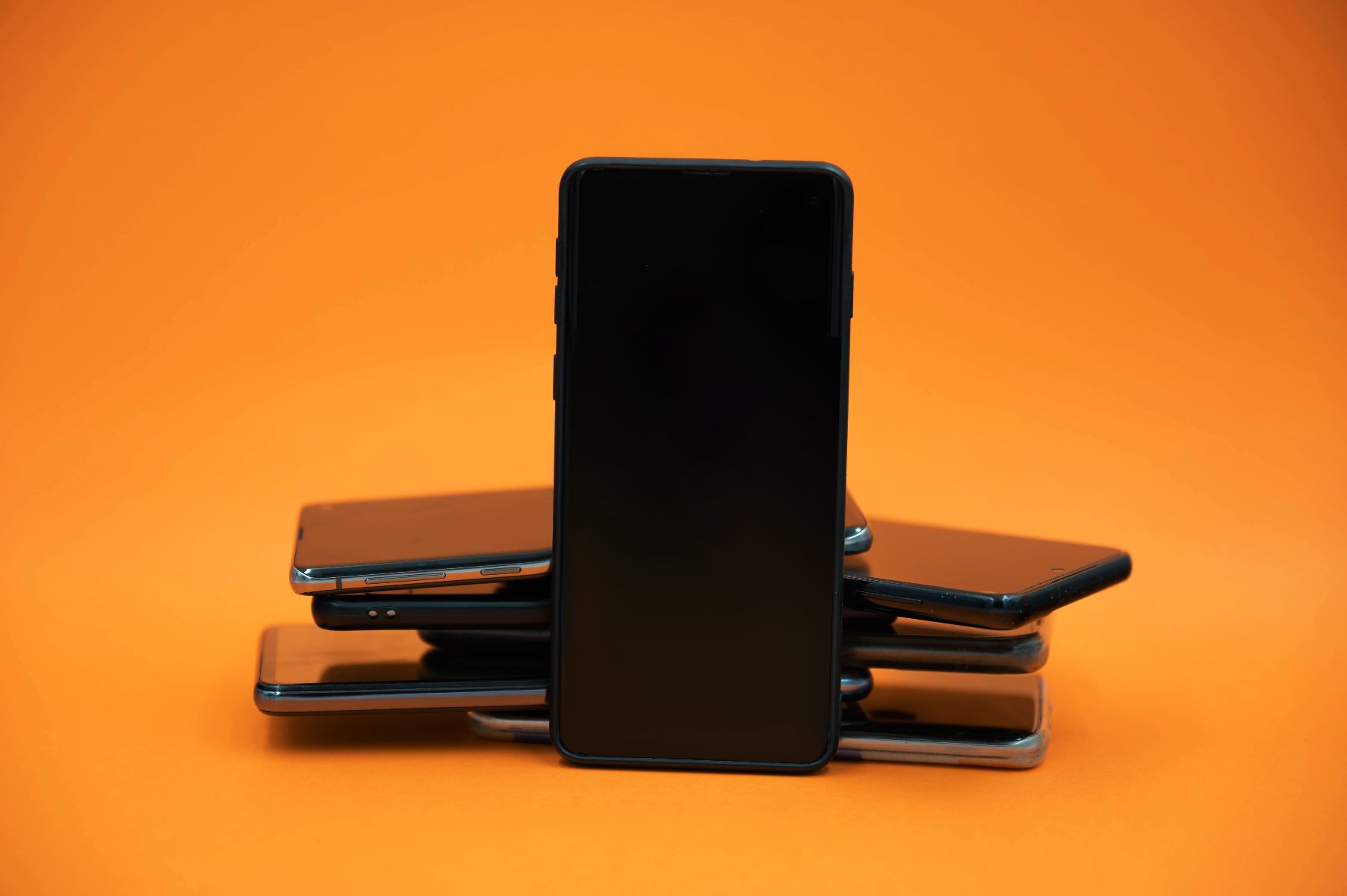 Czarny smartfon stojący pionowo na tle ułożonych poziomo innych smartfonów. Wszystko na pomarańczowym tle. - grafika artykułu