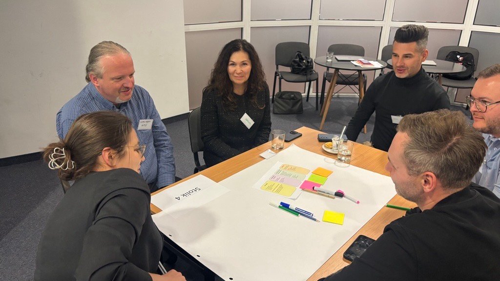 Grupa osób siedząca przy drewnianym stole. Na stole duża biała kartka, małe kolorowe karteczki oraz kolorowe markery. W tle kilka krzeseł i mniejsze stoliki.