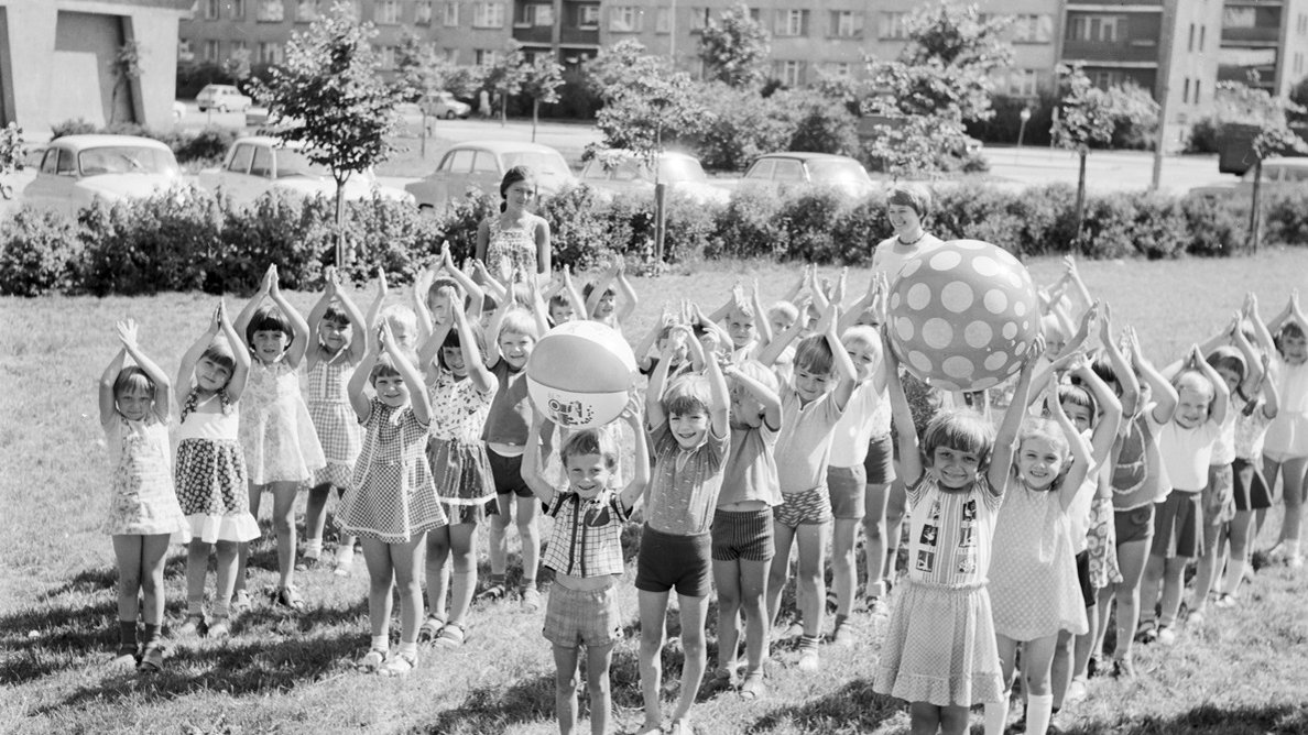 Dzieci stojące na trawie w czterech rzędach z uniesionymi do góry rękoma. Dwoje dzieci trzyma nad głowami duże piłki plażowe. W tle kilka zaparkowanych samochodów, drzewa i bloki mieszkalne. Zdjęcie czarno-białe.