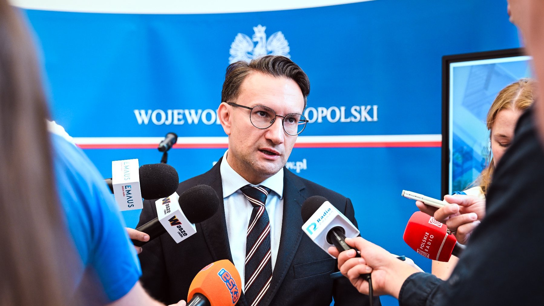 Mężczyzna w garniturze i okularach przemawia do licznych mikrofonów. W tle widać niebieski baner z godłem i napisem "WOJEWODA WIELKOPOLSKI".