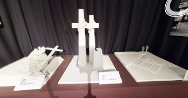 Biała makieta na stole. Przedstawia dwa krzyże obleczone sznurem i mniejszy pomnik, który przypomina orła. Przed makietą stoi tabliczka informacyjna.