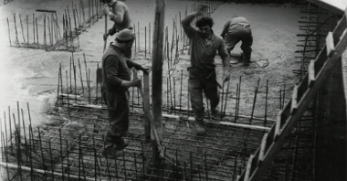 Czarno białe zdjęcie przedstawia pracowników, którzy przygotowują fundamenty pod pomnik. Czterech mężczyzn ubranych w stój roboczy montuje pionowe metalowe drążki.