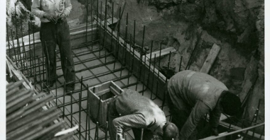 Czarno białe zdjęcie. Dwóch mężczyzn i kobieta ubrani w robocze ubrania przygotowują fundamenty pod pomnik.