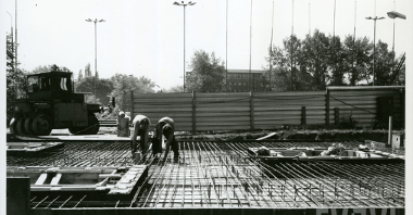 Czarno-białe zdjęcie. Dwóch mężczyzn przygotowuje fundamenty pod pomniki. Za nimi stoi maszyna, która prasuje beton. Teren jest odgrodzony płotem. W oddali widać budynek i drzewa.
