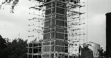 Czarno-białe zdjęcie przedstawia pomnik "Jedność". Wokół jednego z krzyży stoi rusztowanie. Pod rusztowaniem stoi dwóch mężczyzn. W prawym górnym rogu zwisa hak od dźwigu.