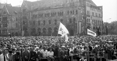 Czarno-białe zdjęcie przedstawia tłum poznaniaków, którzy przyszli oglądać odsłonięcie pomnika. Nad tłumem unoszą się dwie flagi "Solidarość". Za tłumem budynek Filharmonii Poznańskiej.