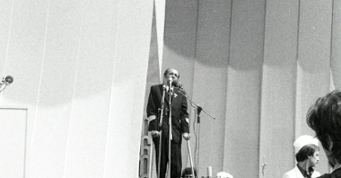 Czarno-biale zdjęcie. Na tle biało-czerwonych płacht na scenie przemawia Stanisław Matyja.