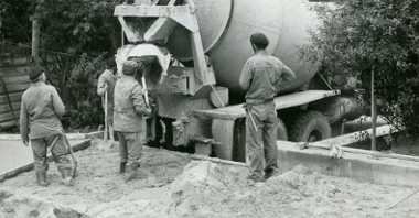 Czarno-białe zdjęcie. Czterech mężczyzn wylewa z betoniarki materiał do fundametu. Mężczyźni są ubrani w stroje robocze i mają w rękach łopaty.