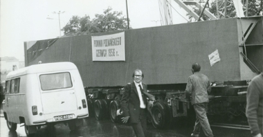 Czarno-białe zdjęcie przedstawia transport jednej z części pomnika. Część leży na przyczepie ciężarówki. Na części wisi płachta z napisem: "Pomnik Poznańskiego Czerwca 1956 r.". Wokół transportu idzie kilka osób.