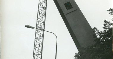 Czarno-białe zdjęcie przedstawia podnoszenie części pomnika przez dźwig. Pomnik jest pochylony nad drzewami w parku.