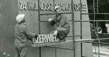 Czarno-biale zdjęcie przedstawia robotników, którzy montują napis na jednej z części pomnika. Mężczyźni stoją na rusztowaniu i trzymają napis: "Czerwiec".
