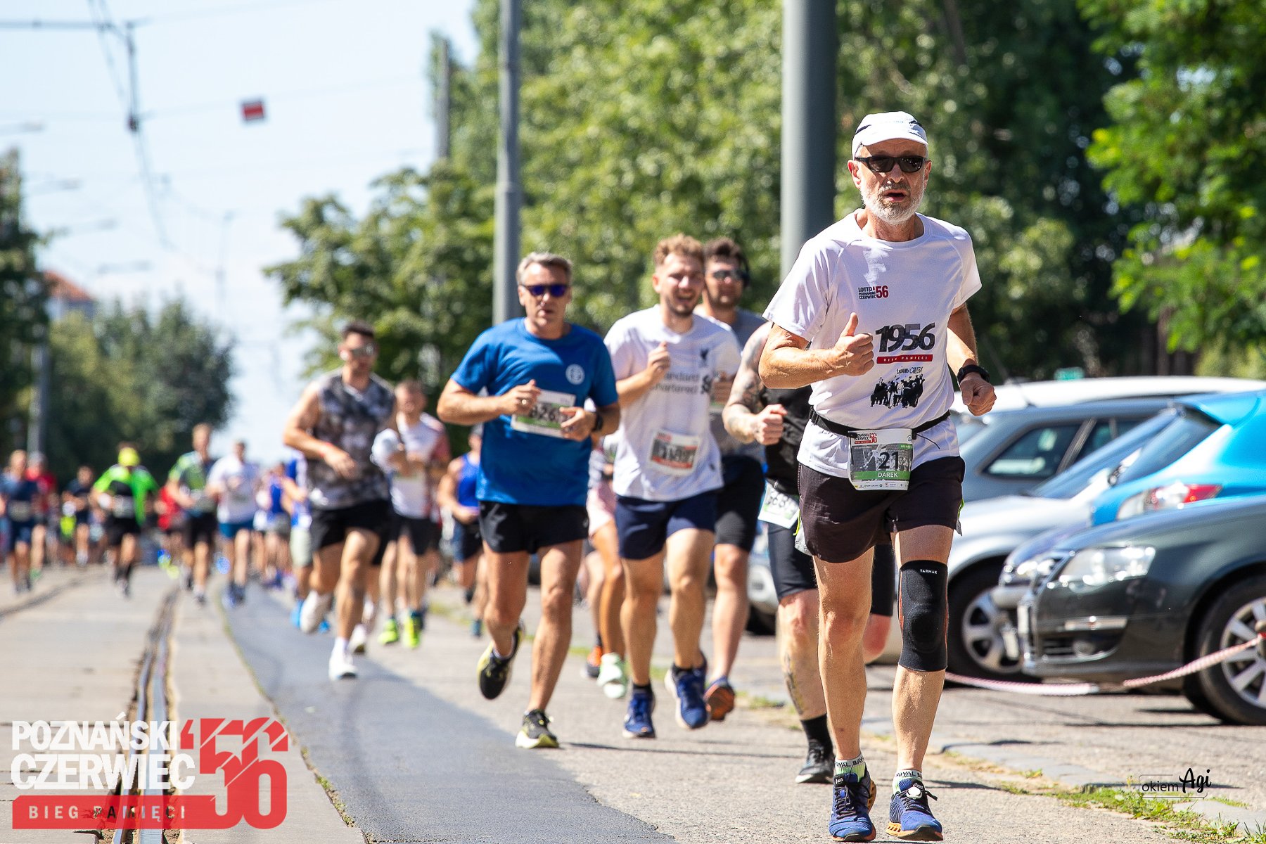 Grupa mężczyzn biegnie w stronę obiektywu. Są w różnym biegu i ubrani w stroje sportowe. W tle samochody, drzewa i reszta biegaczy. - grafika artykułu