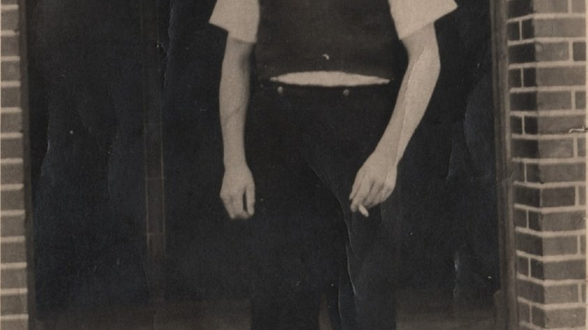 Stare, szare zdjęcie młodego mężczyzny. Stoi w progu wejścia. Ma czarne spodnie, kamizelkę i białą koszulę z krótkim rękawem. W lewej ręce trzyma papierosa. Krzywo uśmiecha się do obiektywu.