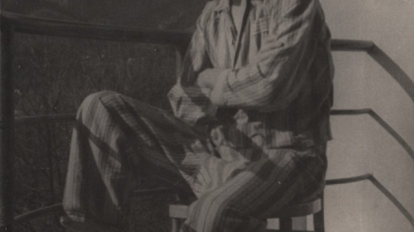 Stare, szare zdjęcie mężczyzny, który siedzi na balkonie na krześle. Ma na sobie "pasiak" czyli strój z obozu koncentracyjnego. Jest odwrócony bokiem do obiektywu, patrzy w dal.