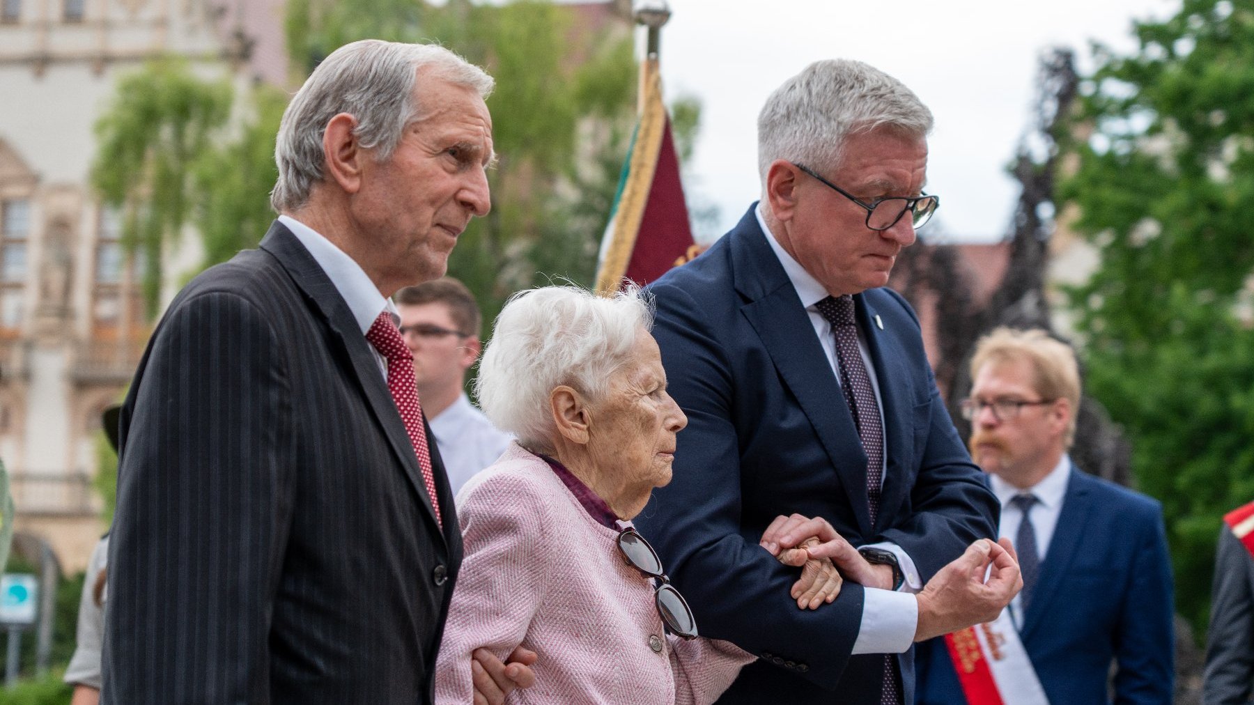 Na zdjeciu dwóch męzczyzn, pomagają iść starszej kobiecie