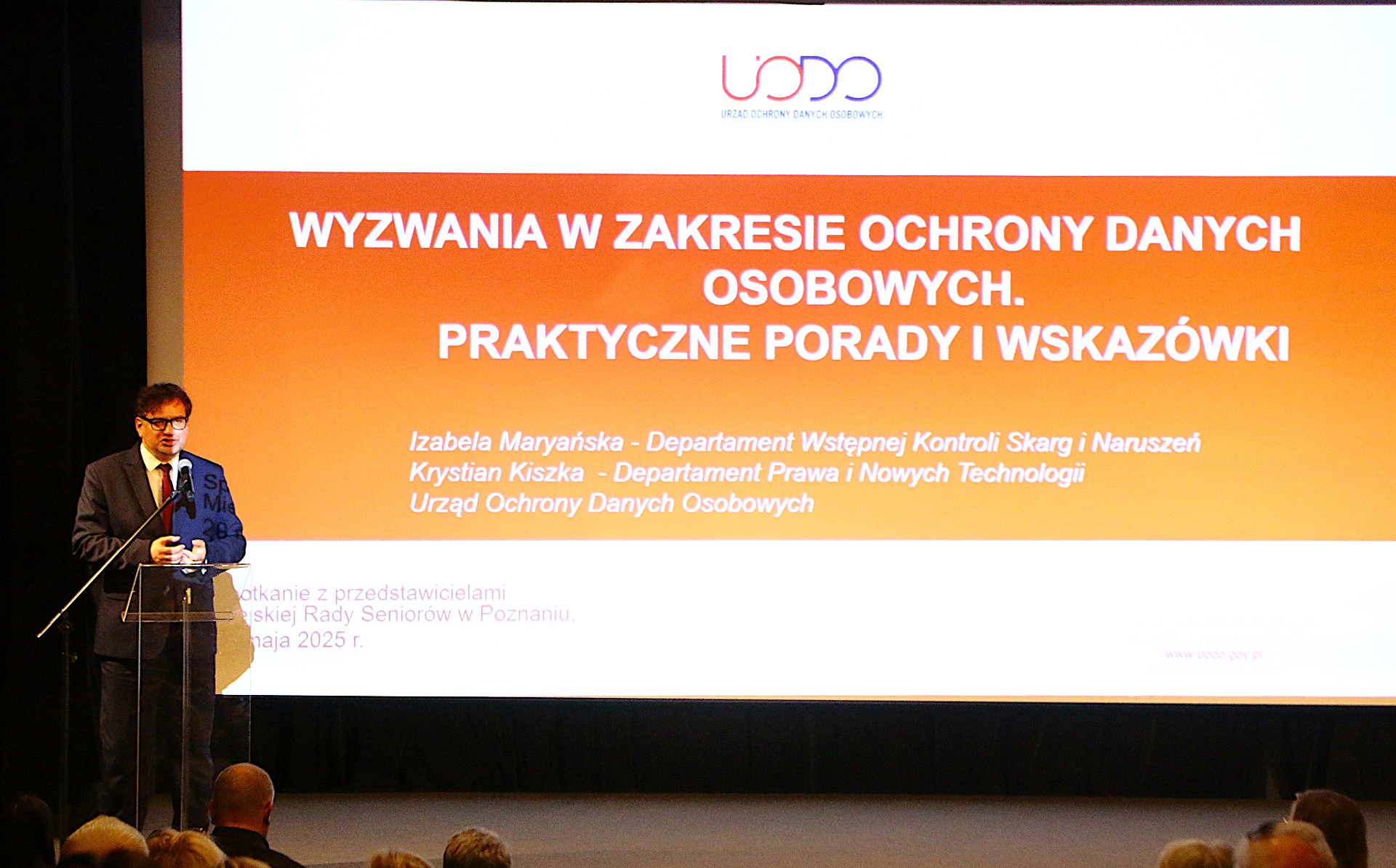 Na zdjęciu widzimy Pana prelegenta na tle slajdu z prezentacji omawianej na spotkaniu. Slajd w kolorze pomarańczowym. - grafika artykułu