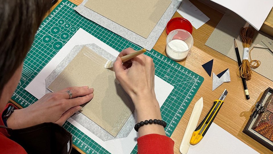 Na zdjęciu widoczne są dłonie, które wyklejają kartki na notes, widoczne są miarki, ołówki