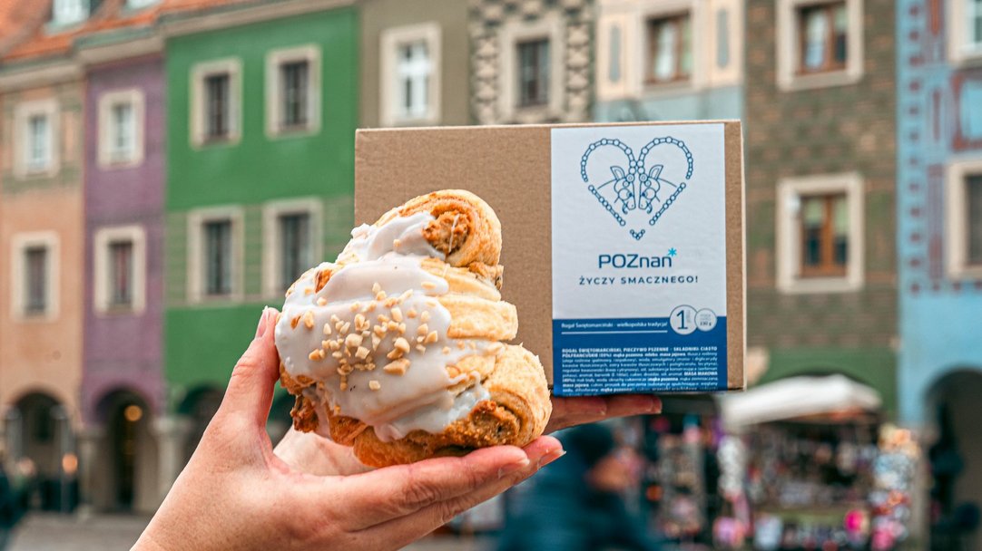 Rogal świętomarciński na tle kamieniczek na Starym Rynku. Za rogalem kartonik a na nim napis Poznań życzy smacznego.