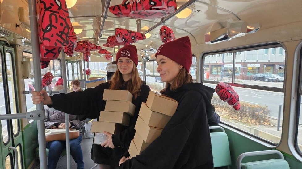 Wnętrze trmawaju a w nim dziewczyny w czerwonych czapkach z napisem Czyń dobro. Dziewczyny trzymają w rękach kartony z rogalami.