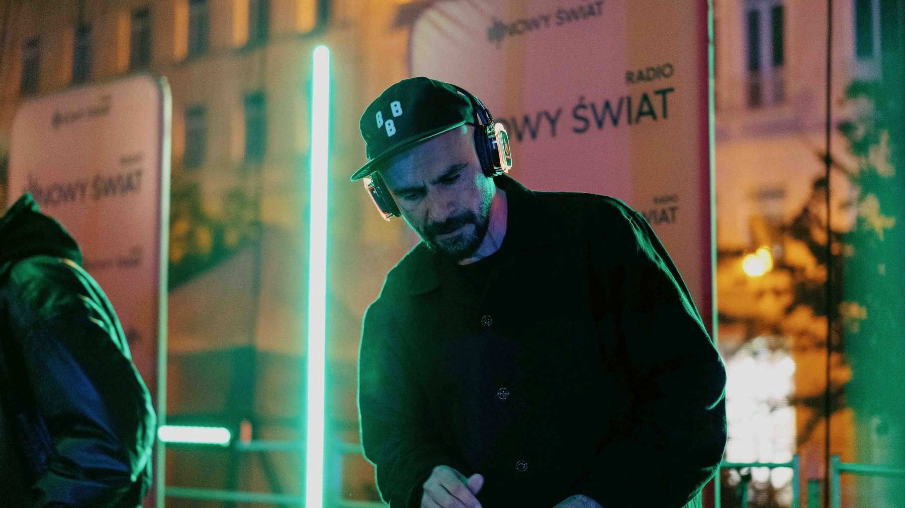 Mężczyzna ze słuchawkami na głowie stoi przy konsoli DJ'a. Za nim stoją banery z napisami "Radio Nowy Świat".