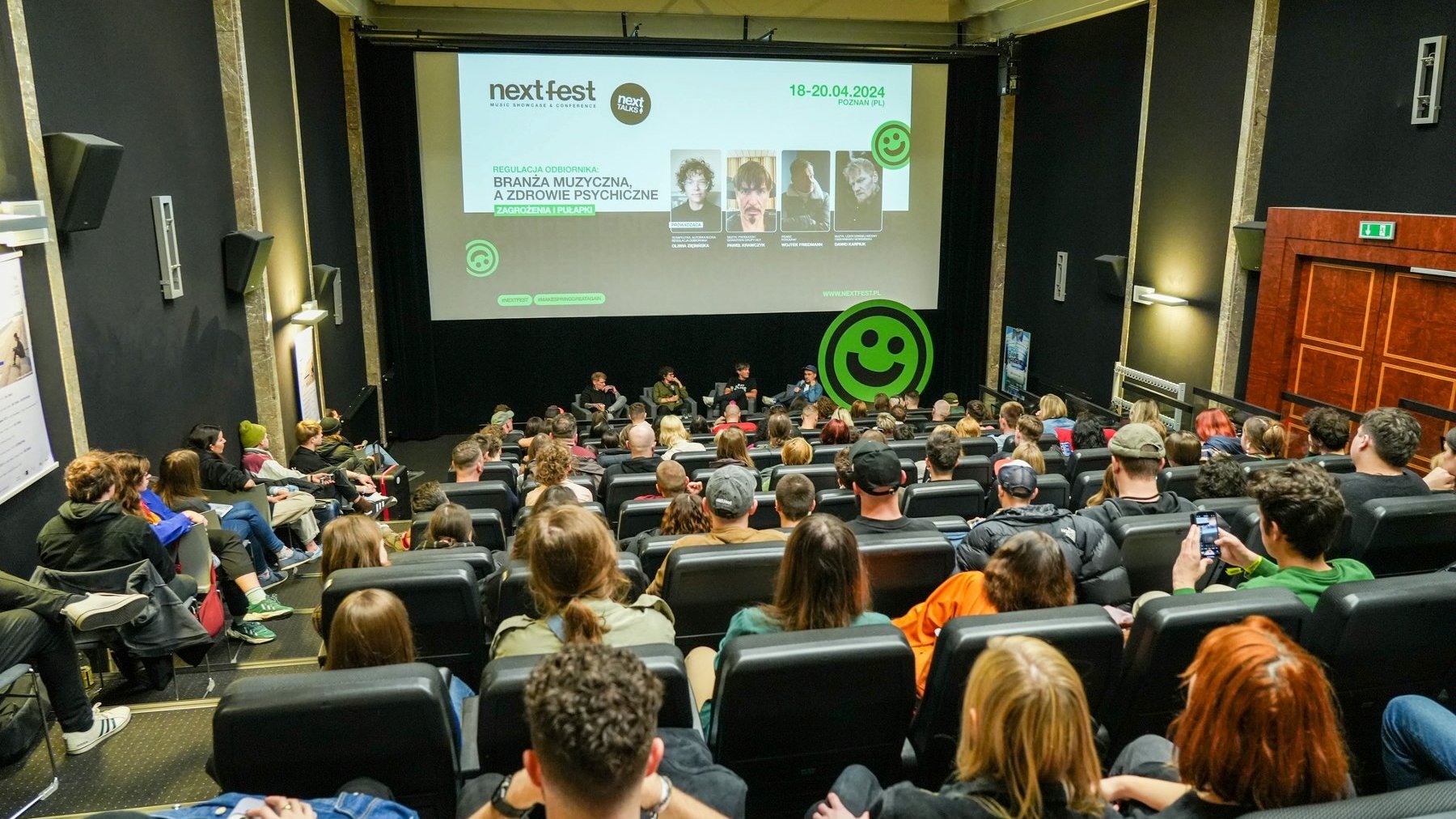 Sala konferencyjna. Kadr od tyłu publiczności. Słuchacze biorą udział w dyskusji na temat zdrowia psychicznego. Na środku sceny siedzi czterech mężczyzn. Za nimi duży ekran wyświetla tytuł dyskusji.