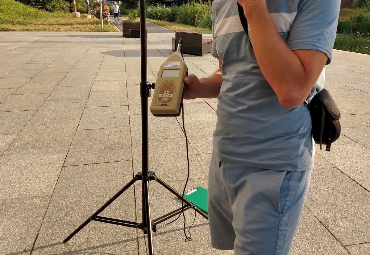 osoba stojąca i trzymająca urządzenie do pomiaru hałasu, nie widać jej twrazy w kadrze - grafika artykułu