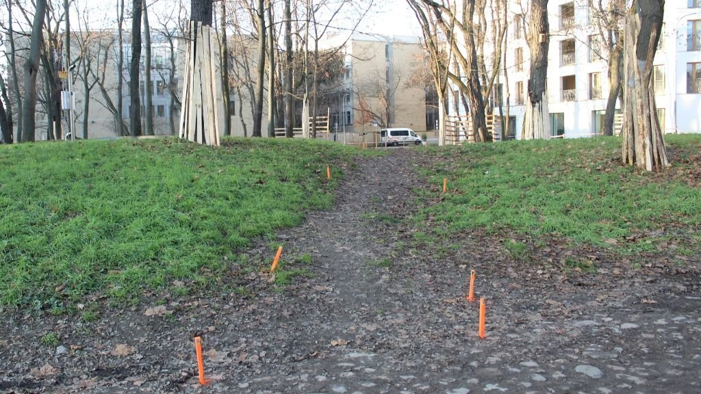 Piaszczysto-trawiasta ścieżka na lekkim wzniesieniu. Po bokach widoczne są pomarańczowe znaczniki wbijane w ziemię. Na wzniesieniu w oddali widoczna jest biała furgonetka i ogrodzenie z drewnianych pali. W tle widoczne są bloki mieszkalne z białymi fasadami i gołe drzewa.