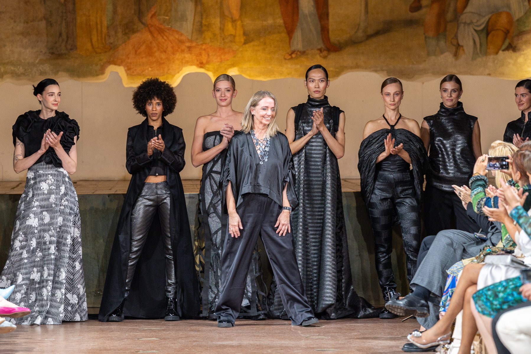Projektantka stoi na wybiegu wśród modelek w czarnych kreacjach, odbierając brawa po pokazie mody.