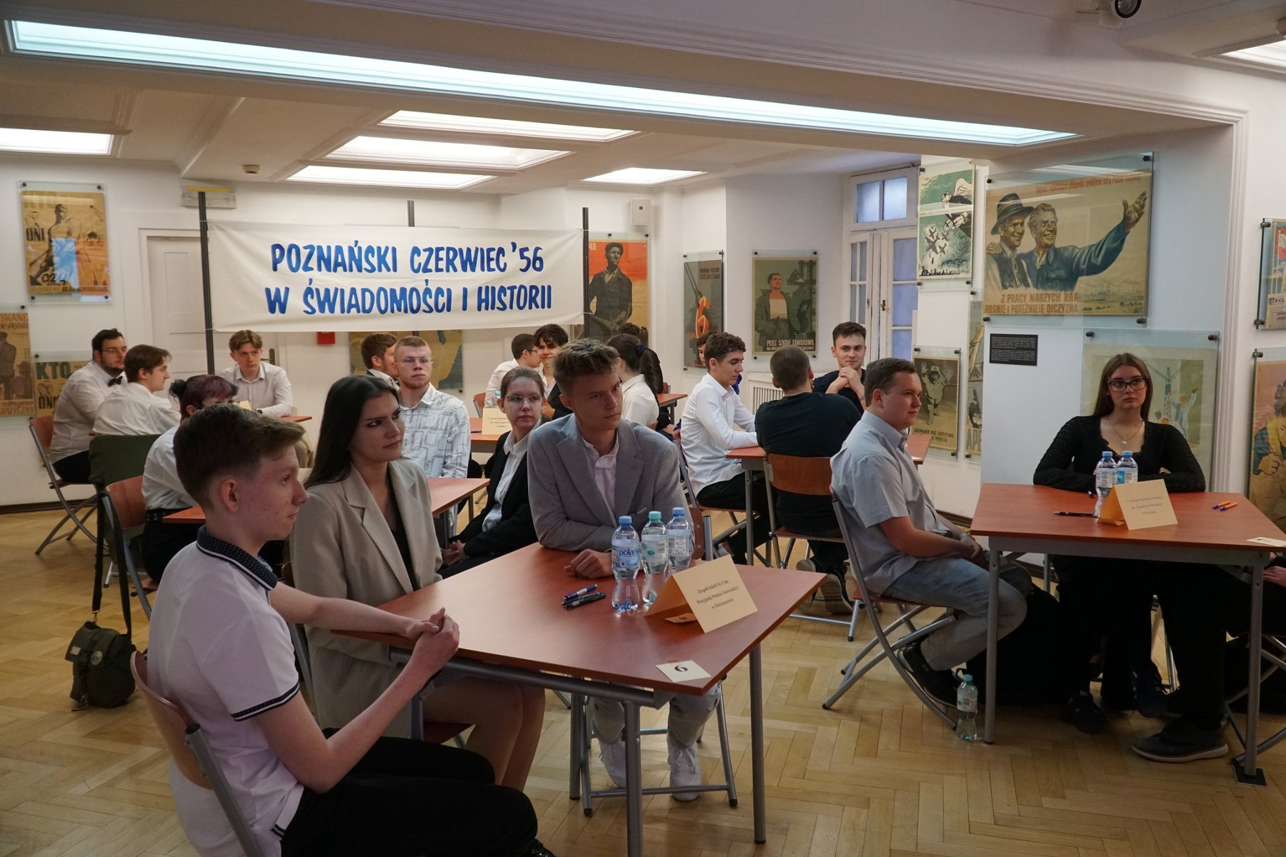 W poniedziałek 3 czerwca odbył się finał VII Testu Wiedzy o Poznańskim Czerwcu / fot. WMN Przy pięciu stolikach siedzą 3-osobowe druzyny uczniów. Są ubrani na galowo. Z tyłu biały transparent z niebieskim napisem 'Poznański Czerwiec '56 w świadomości i historii". Uczniowie siedzą w sali ozdobionej obrazami z okresu PRL-u.