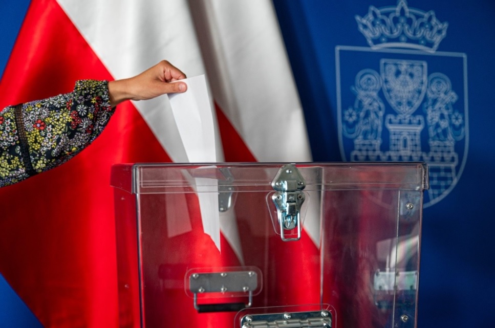 ręka wrzuca kartę do przezroczystej urny wyborczej, w tle flagi z herbem Poznania