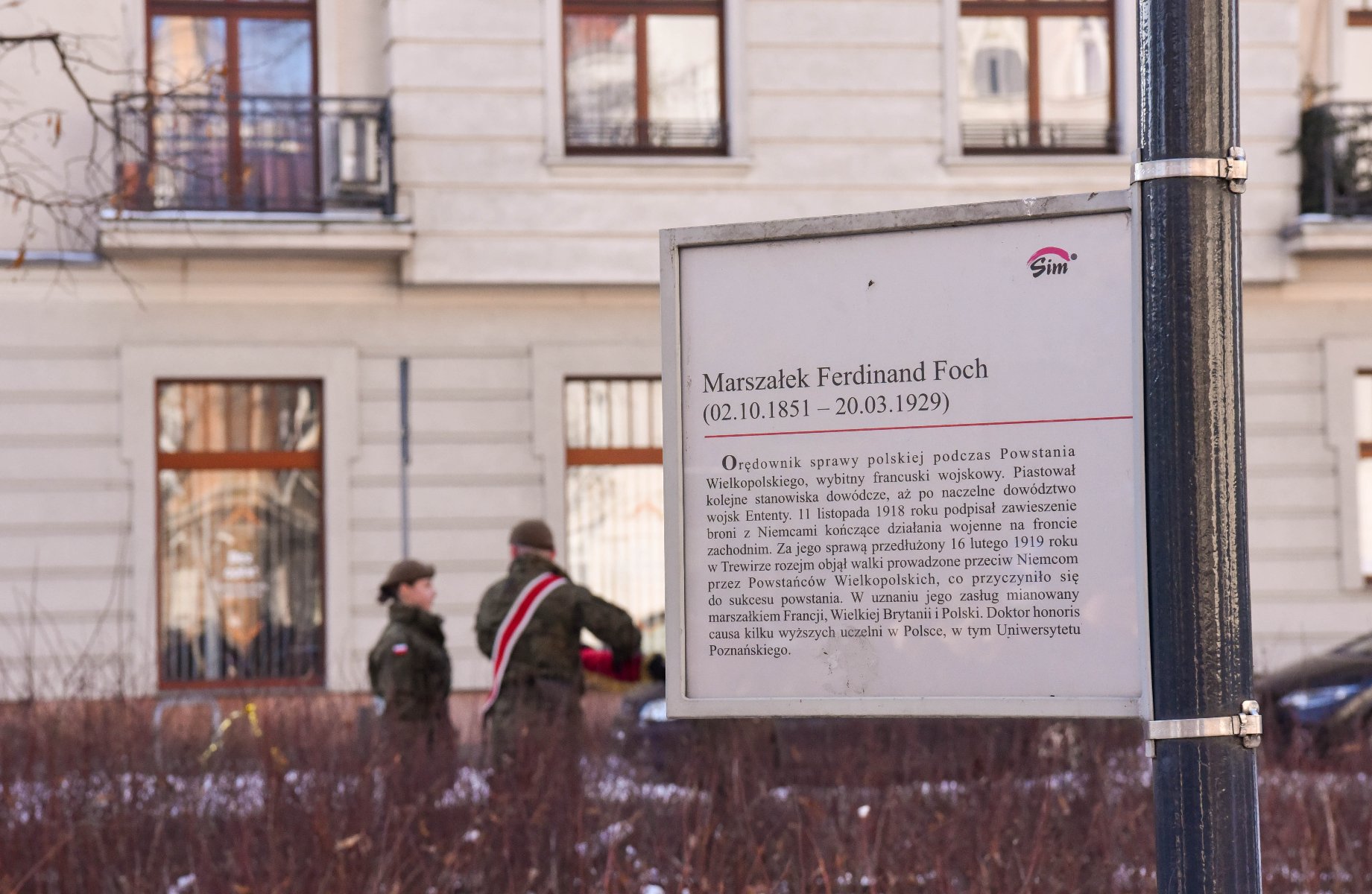 Tabliczka z dedykacją dla Marszałka Ferdinanda Focha, opisująca jego zasługi i znaczenie. Tabliczka jest w języku polskim i zawiera tekst: "Marszałek Ferdinand Foch (02.10.1851 - 20.03.1929) Ordownik sprawy polskiej podczas Powstania Wielkopolskiego, wybitny francuski wojskowy... (ciąg dalszy)." W tle znajdują się dwie osoby w mundurach wojskowych, jedna z nich ma na ramieniu biało-czerwoną szarfę.