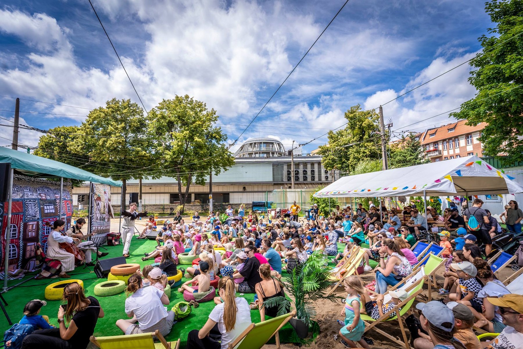 Lato na Madalinie świętuje już 10 lat! Organizatorzy przygotowali coś specjalnego na rozpoczęcie tegorocznych wakacji. Tłum dzieci i dorosłych siedzi na plażowych leżakach przez sceną. Na scenie dwuosobowy zespół, kobieta gra na wiolonczeli a mężczyzna na gitarze. Za nimi dekoracje. W tle budynki i wysokie, zielone drzewa. Niebo jest blękitne i pokryte białymi chmurami.