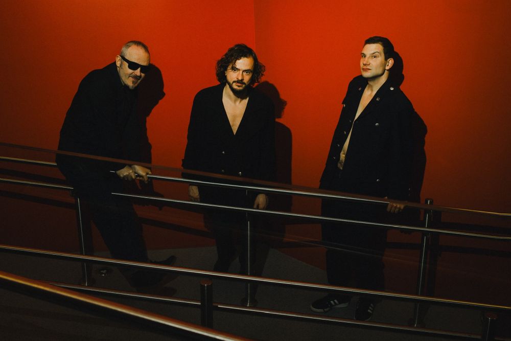 Trzech muzyków pozuje na tle czerwonej ściany.