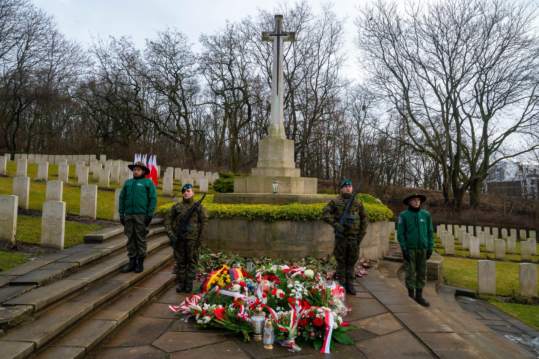 Uroczystość na cmentarzu wojennym. W centralnym punkcie znajduje się wysoki, kamienny krzyż z metalowym mieczem na postumencie, przed którym złożono liczne wieńce i zapalono znicze. Po obu stronach pomnika pełniona jest warta honorowa: stoją dwaj żołnierze w pełnym umundurowaniu z bronią oraz dwie harcerki w zielonych mundurach. W tle widoczne są rzędy jednakowych, jasnych tablic nagrobnych na trawiastym zboczu oraz bezlistne drzewa.