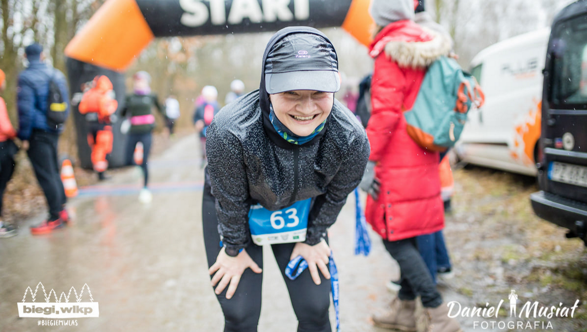 Uśmiechnięta uczestniczka biegu, w czapce i z numerem startowym, w tle brama z napisem "start" oraz inne osoby, których postacie są rozmazane.