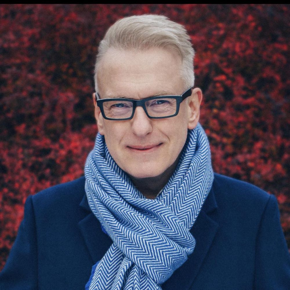 Mariusz Szczygieł fot. Damian Maliszewski Uśmiechnięty mężczyzna w szaliku na tle czerwonego, jesiennego bluszczu.