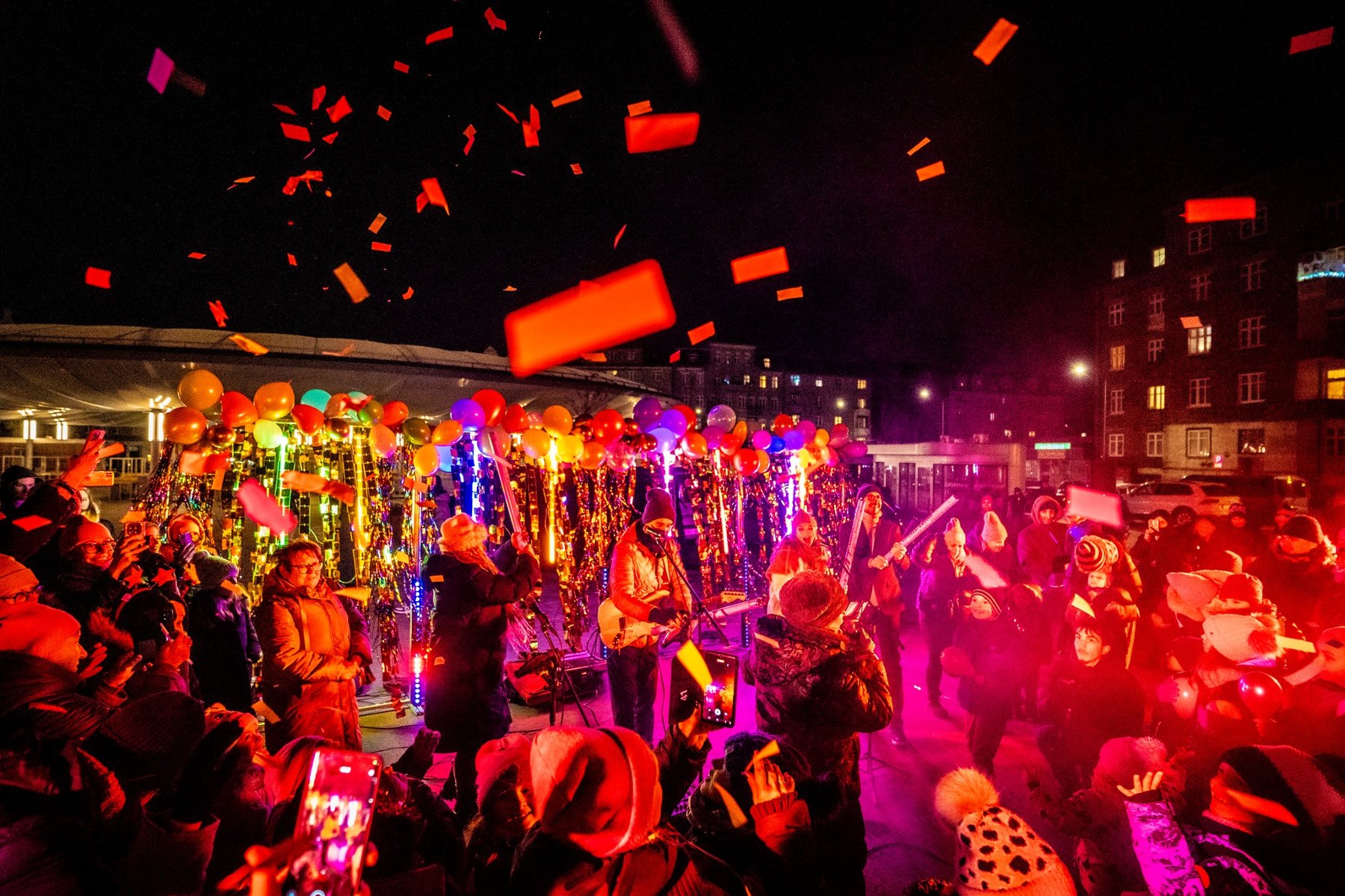 Widok na bawiący się tłum. W tle widać balony i kolorowe iluminacje. Widać jak na ludzi opada konfetti. Wydarzenie ma miejsce gdy już jest ciemno na zewnątrz.