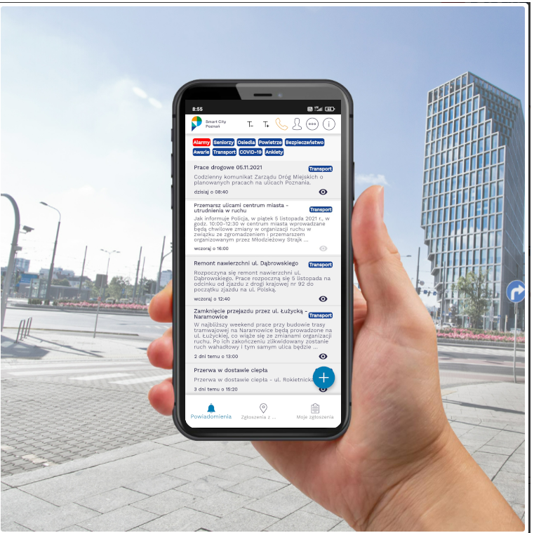 Zdjęcie przedstawia dłoń trzymającą smartfon z działającą aplikacją Poznań Smart City. Aplikacja wyświetla widok powiadomień. W tle widoczny jest budynek wieżowca Bałtyk, znajdującego się w Poznaniu, obok Ronda Kaponiera.