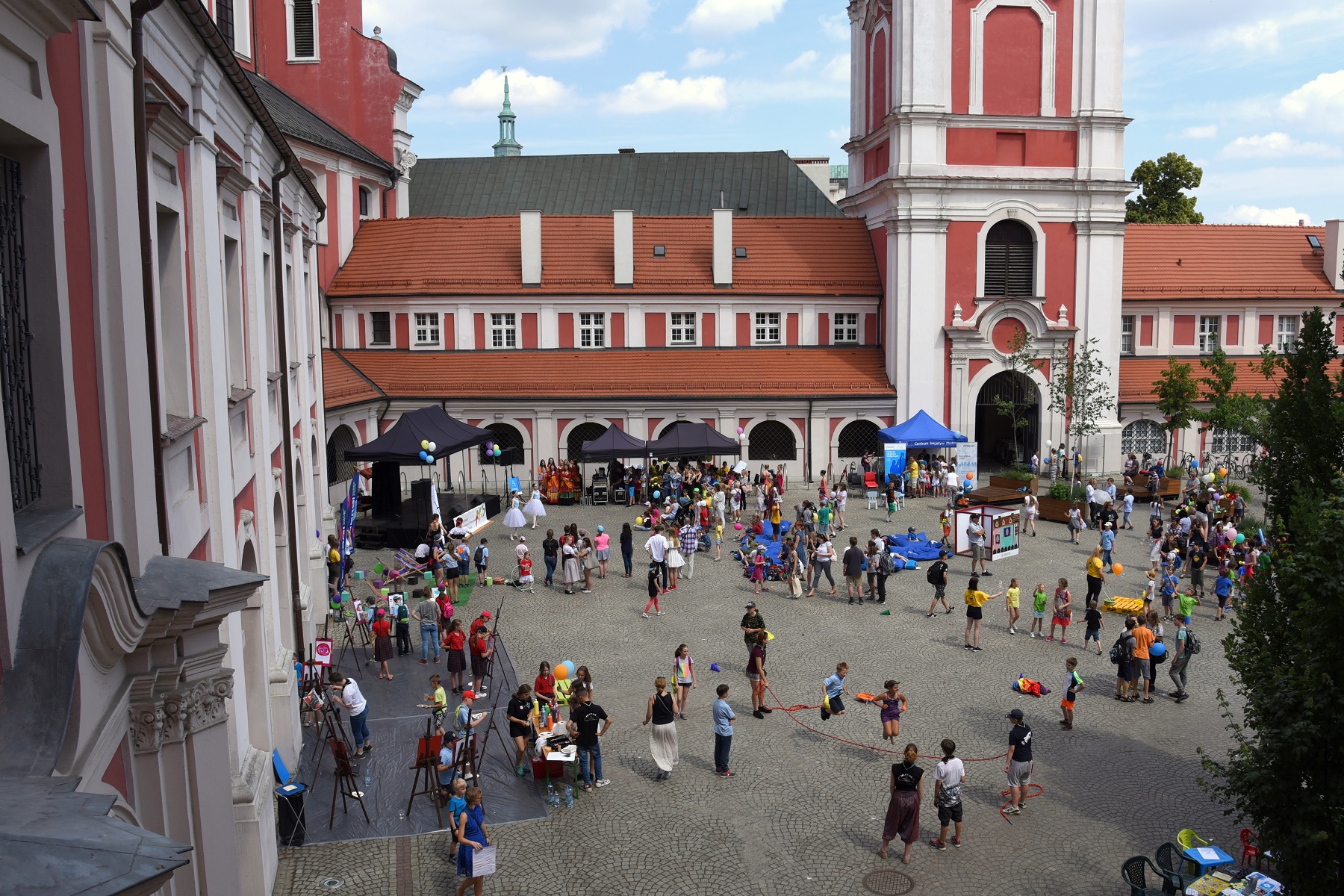 24 i 26 czerwca odbędą się warsztaty metodą World Café, które mają wesprzeć opracowanie "Planu działań na rzecz dzieci i młodzieży w Poznaniu" Widok z góry na dziedziniec urzędu miasta podczas wydarzenia plenerowego. Wokół stoją budynki z czerwonymi dachami i arkadami. Na brukowanym placu znajduje się wiele osób, namioty i stragany, a po lewej stronie widoczne są sztalugi malarskie.
