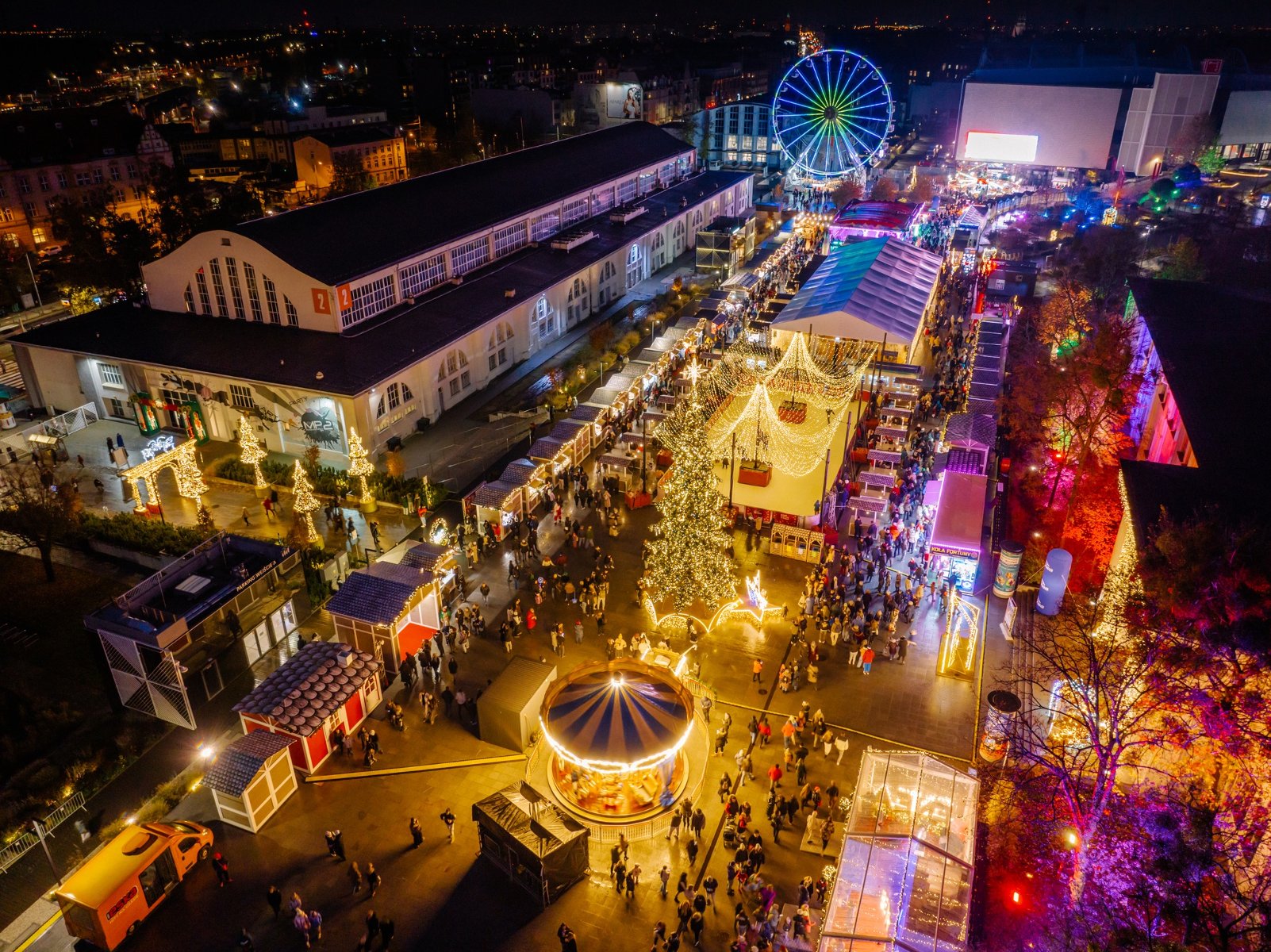 Widok z lotu ptaka na Jarmark Świąteczny na Międzynarodowych Targach Poznańskich. Na dziedzińcu stoją budki, karuzela wiedeńska i lodowisko. Między atrakcjami poruszają się tłumy ludzi. W tle wysoki diabelski młyn, który świeci na różne kolory. W oddali panorama miasta i inne budynki na terenie MTP.