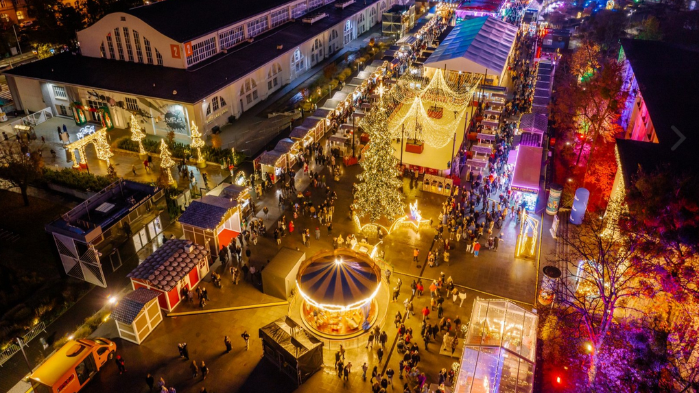 Widok z lotu ptaka na Jarmark Świąteczny na Międzynarodowych Targach Poznańskich. Na dziedzińcu stoją budki, karuzela wiedeńska i lodowisko. Między atrakcjami poruszają się tłumy ludzi. W tle wysoki diabelski młyn, który świeci na różne kolory. W oddali panorama miasta i inne budynki na terenie MTP.